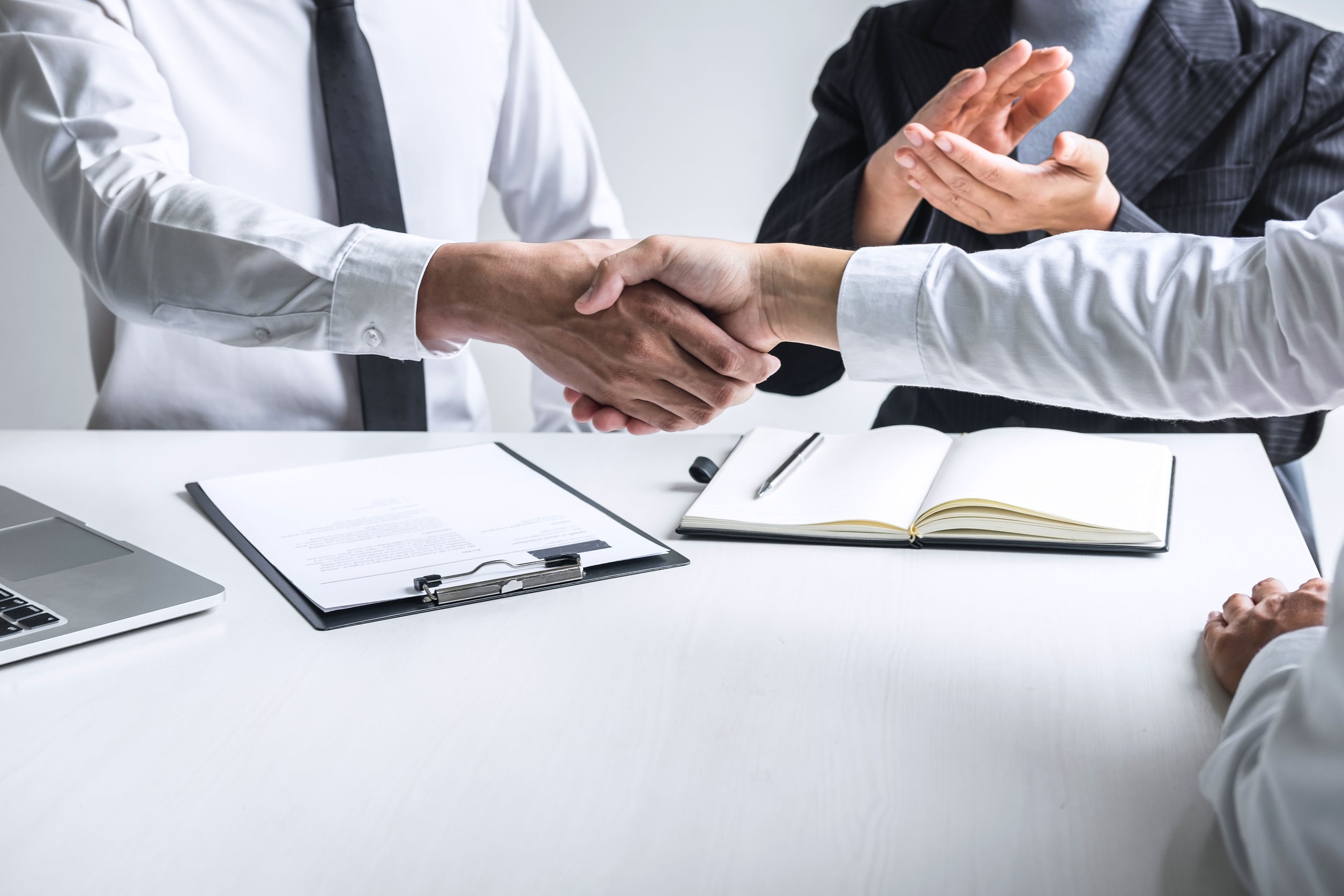 Two people sitting at the table shake hands with each other. One sitting in the middle clap his hands.